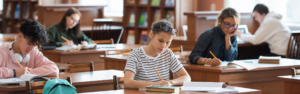Students in a library study hall: a girl in a striped shirt writes at a desk with books open and glasses on her head; others study in the background.