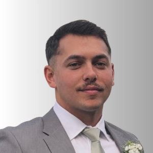 Man in a gray suit, white shirt, and pale tie with a boutonniere on the lapel; partial portrait cropped at the top of the head.
