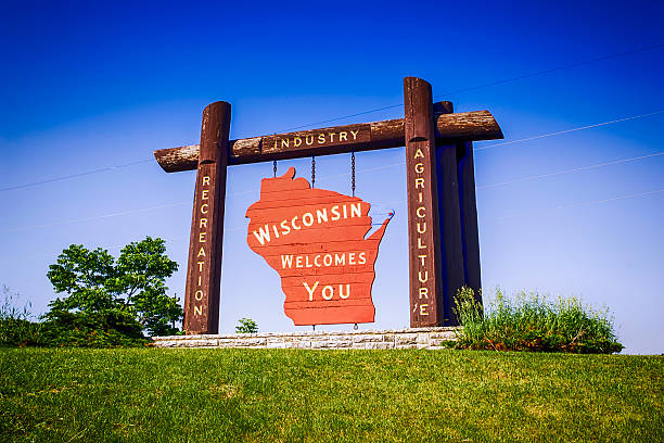 Rustic wooden gateway sign in Wisconsin: orange Wisconsin silhouette with 'Wisconsin welcomes you' on a grassy hill under a blue sky.