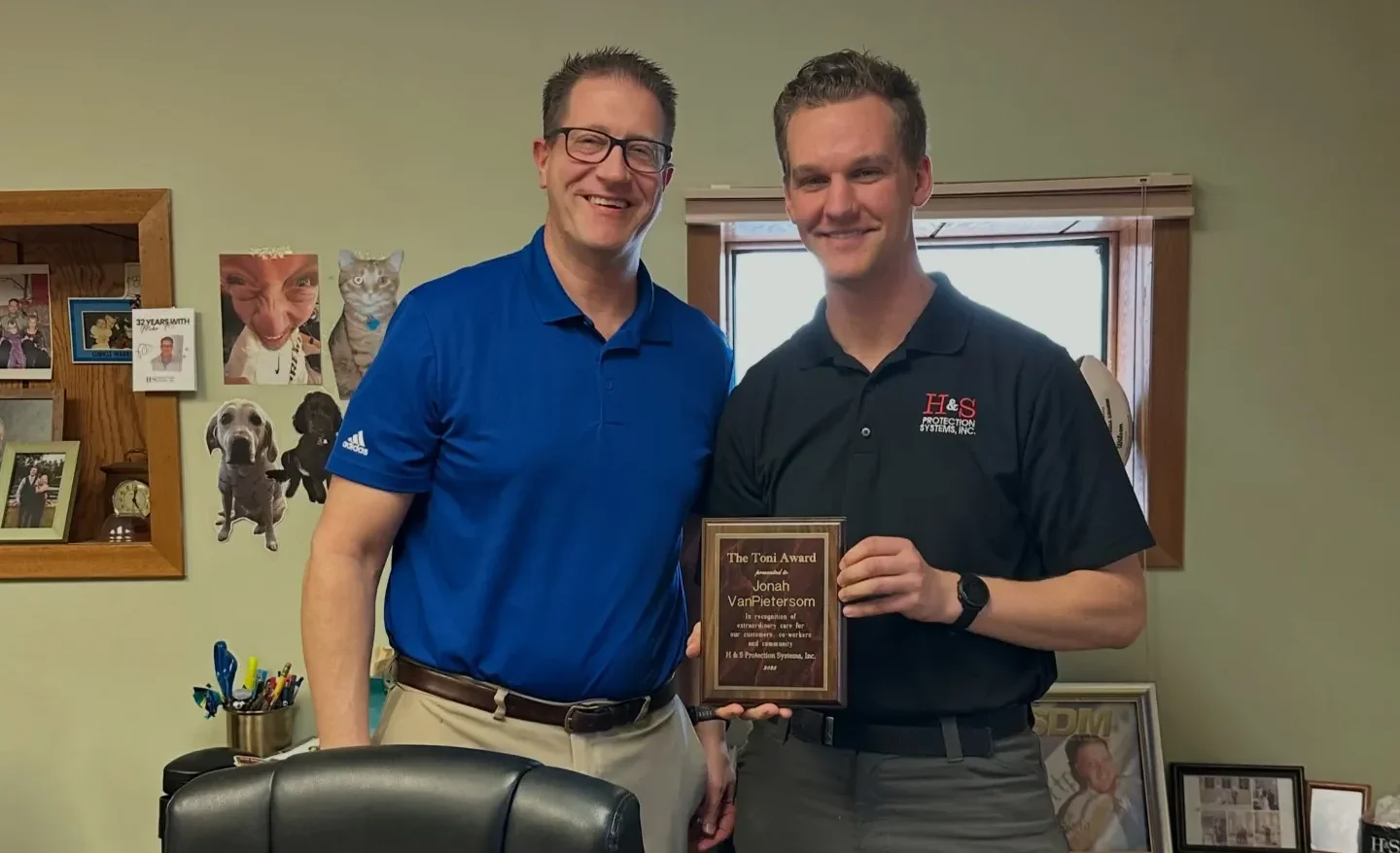 Two men stand in a home office, smiling, holding a wooden award plaque between them.