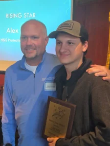 Two men pose for a photo at an award event; the man on the left wears a light blue jacket, the man on the right in a cap smiles while holding a Rising Star Award plaque. In the background a screen reads 'RISING STAR' and 'Alex'.