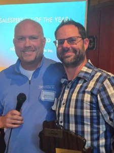Two men smile for a selfie at a conference; one holds a microphone and wears a blue polo with a name badge in front of a blue event screen.