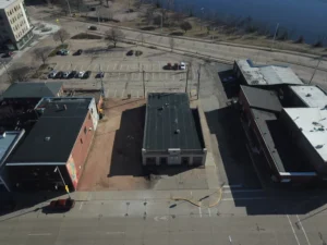 Aerial view of a small downtown with a central white flat-roofed building surrounded by parking lots and brick structures, with a river in the background.