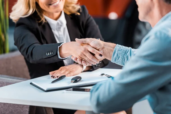 Two professionals shake hands across a round table with pens and a notebook, signaling an agreement.